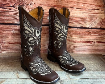 May include: A pair of brown leather cowboy boots with intricate white embroidery featuring a butterfly design. The boots have a pointed toe and a small heel.