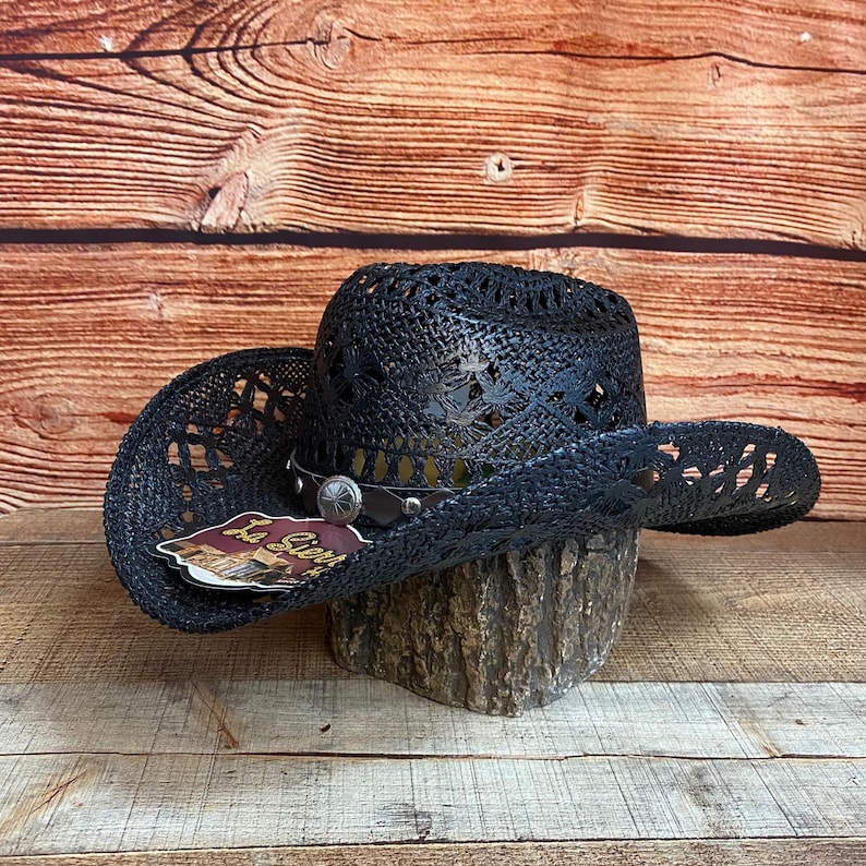 May include: Black straw cowboy hat with a decorative band and a cutout design. The hat is sitting on a wooden surface.