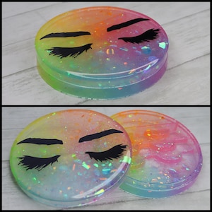 Puede incluir: Dos posavasos redondos de resina de color arcoíris con un diseño de ojo y ceja negro. Los posavasos están llenos de purpurina iridiscente.
