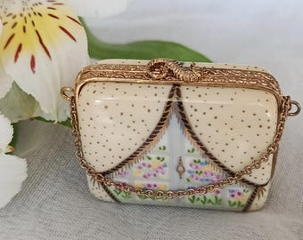 Könnte beinhalten: Kleine, rechteckige, cremefarbene Schmuckschatulle mit goldfarbener Kette und Verzierungen. Die Schatulle zeigt eine handgemalte Fensterszene mit bunten Blumen und einem gepunkteten Muster. Die Schatulle ist wahrscheinlich aus Porzellan oder einem ähnlichen Material.