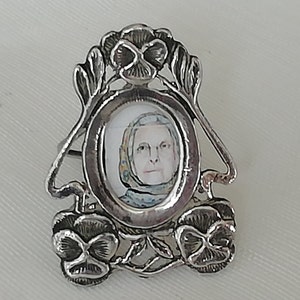 Puede incluir: Broche plateado con un retrato ovalado de una mujer con pañuelo en la cabeza. El broche presenta un diseño floral elaborado con tres flores similares a pensamientos. El retrato está centrado en el marco del broche.