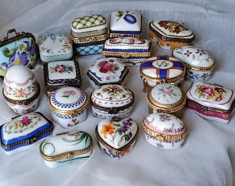 May include: A collection of decorative porcelain trinket boxes in various shapes, including oval, square, and heart-shaped. The boxes feature floral designs, gold trim, and intricate details. Some have hinged lids and are set against a white background.