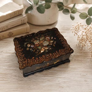 May include: A rectangular black lacquer box with a scalloped edge, featuring an intricate floral inlay made of iridescent mother-of-pearl and gold-toned painted details along the border and the centre of the lid, resting on four small feet.