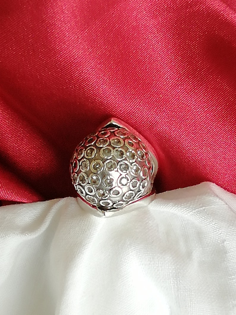 May include: A silver ring with a round, textured design. The ring is set with small, clear stones.