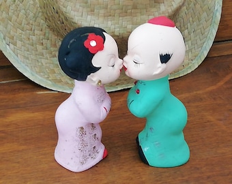 May include: A pair of ceramic figurines of a man and woman in traditional clothing kissing. The woman is wearing a pink dress with gold accents and a red flower in her hair. The man is wearing a teal green robe with gold accents.