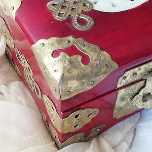May include: A red wooden box with ornate brass corner protectors and a clasp. The box features decorative brass accents and a white carved design on the lid. The box is likely used for storing jewelry or other small items.