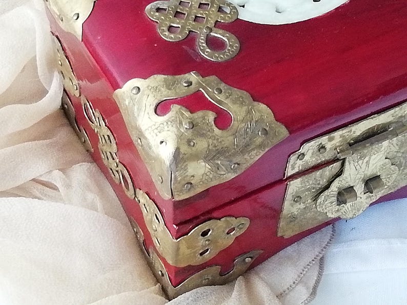 May include: A red wooden box with ornate brass corner protectors and a clasp. The box features decorative brass accents and a white carved design on the lid. The box is likely used for storing jewelry or other small items.