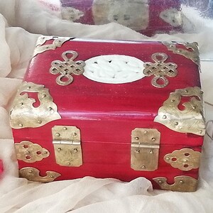 May include: A red wooden box with brass hardware and decorative accents. The box features a white oval center and ornate corner pieces. The box is likely used for storage or display.