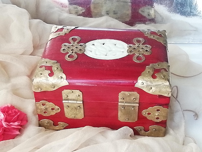 May include: A red wooden box with brass hardware and decorative accents. The box features a white oval center and ornate corner pieces. The box is likely used for storage or display.