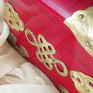 May include: Close-up of a red lacquered wooden box with ornate brass hardware. The box features decorative brass accents in various shapes, including cloud-like designs and a central knot motif. The background includes a soft, off-white fabric.