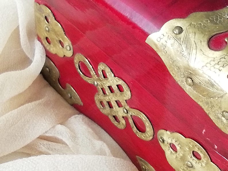 May include: Close-up of a red lacquered wooden box with ornate brass hardware. The box features decorative brass accents in various shapes, including cloud-like designs and a central knot motif. The background includes a soft, off-white fabric.