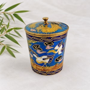 Pode incluir: Um recipiente cilíndrico de metal com tampa, apresentando esmalte azul vibrante decorado com padrões de fio dourado, motivos de grous brancos e pretos e nuvens estilizadas. A tampa tem um pequeno botão central e um design de dragão.