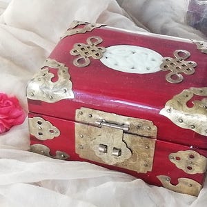May include: A red wooden box with ornate brass corner accents and a brass clasp. The box features a white oval inlay and decorative brass embellishments. A pink rose rests nearby.