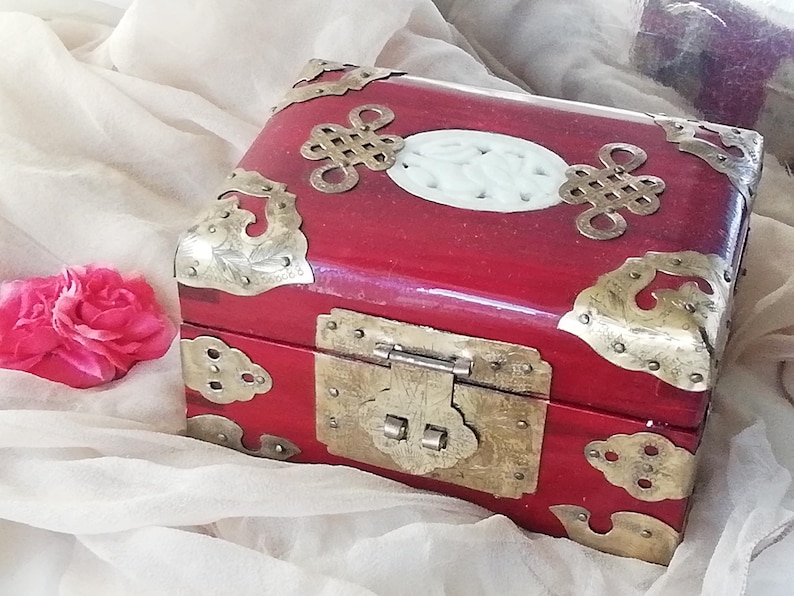 May include: A red wooden box with ornate brass corner accents and a brass clasp. The box features a white oval inlay and decorative brass embellishments. A pink rose rests nearby.