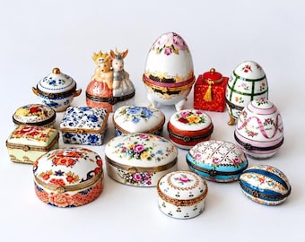 May include: A collection of various porcelain trinket boxes in oval, round, square, and egg shapes. The boxes feature intricate hand-painted floral patterns, gold-tone metal hinges, and decorative motifs including rabbits and ribbons.