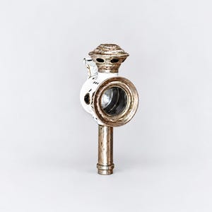 May include: A vintage carriage lamp featuring a white enamelled metal body with a circular glass lens. The lamp has a tarnished silver-toned metal top vent and a matching vertical cylindrical handle extending from the base, showing signs of age.