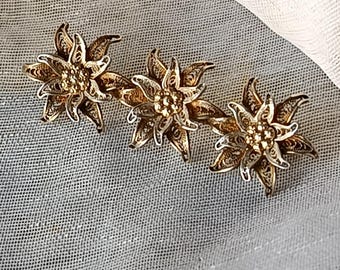May include: A gold-toned metal brooch featuring three connected edelweiss flower shapes. Each flower is crafted with intricate filigree wire petals and a textured, beaded center. The metal has a warm, metallic finish with detailed openwork.
