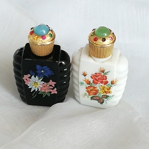 May include: Two small glass perfume bottles, one black and one white, featuring ribbed sides and floral decals. Each bottle has a gold-toned cap topped with a central cabochon stone surrounded by smaller colorful rhinestones.