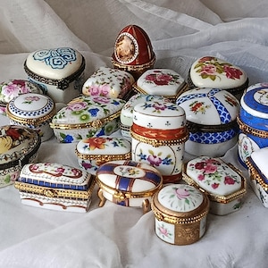 Könnte beinhalten: Eine Sammlung dekorativer Porzellan-Schmuckdosen in verschiedenen Formen, darunter oval, herzförmig und rechteckig. Die Dosen weisen florale und geometrische Designs mit goldfarbenen Verzierungen auf. Die Farbpalette umfasst Weiß, Blau, Rot und Rosa.