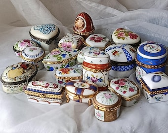 May include: A collection of decorative porcelain trinket boxes in various shapes, including oval, heart, and rectangular. The boxes feature floral and geometric designs, with gold-colored trim. The color palette includes white, blue, red, and pink.