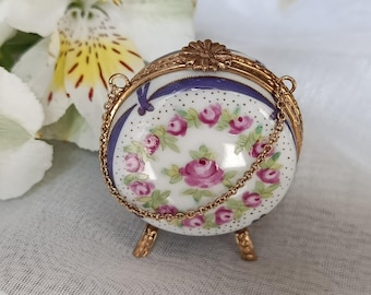 May include: A small, round, porcelain trinket box with a gold-tone metal frame and chain. The box is decorated with pink roses and green leaves on a white background, accented with a blue ribbon design. The box rests on three small gold-tone feet.