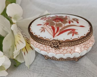 May include: An oval porcelain trinket box with a floral design in red and gold. The box has a gold-colored metal frame and feet, and a pink and white geometric pattern on the sides. The lid features a detailed floral motif.