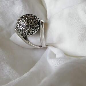 May include: A silver ring with a textured, circular design. The ring has a unique, angular band.