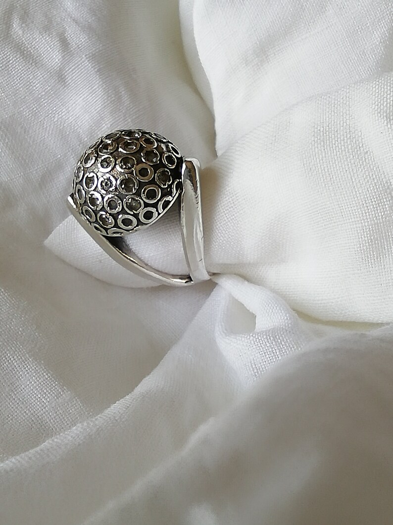 May include: A silver ring with a textured, circular design. The ring has a unique, angular band.