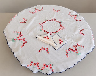Op de afbeelding: Een rond wit tafelkleed met een geschulpte donkerblauwe rand, voorzien van geborduurde rode bloemen en groene stelen. Een bijpassende set gevouwen witte servetten met identiek rood bloemenborduurwerk en donkerblauwe geschulpte randen ligt erbovenop.
