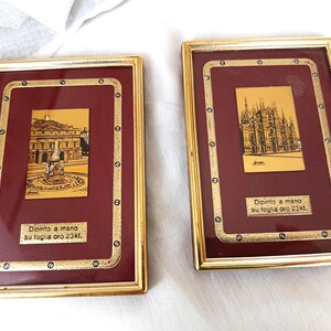 Può includere: Due opere d'arte incorniciate in foglia d'oro. Ognuna presenta un'immagine dorata su sfondo bordeaux, con testo in basso. Le cornici dorate hanno rivetti decorativi. Una raffigura un edificio con una fontana, l'altra una cattedrale.
