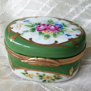 May include: A green and white oval trinket box with a floral design and gold accents. The lid features a delicate floral pattern with pink, white, and yellow flowers. The box is decorated with gold trim and a gold feather design.
