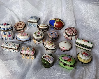May include: A collection of fifteen small porcelain trinket boxes in various shapes including oval, round, rectangular, and egg-shaped. Each box features intricate hand-painted floral patterns, geometric designs, or Chinese characters with metal hinges.