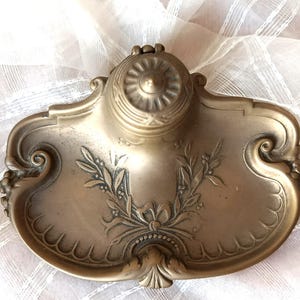 May include: Antique brass inkwell with ornate floral and leaf detailing. The inkwell features a central circular well and a decorative tray. The design includes a laurel wreath and scrollwork, showcasing a vintage aesthetic.