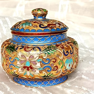 Puede incluir: Un tarro decorativo con tapa y diseños intrincados de flores y volutas. El tarro presenta un fondo dorado con acentos de esmalte de colores en azul, rojo, verde y blanco. La tapa tiene un pequeño pomo redondo.