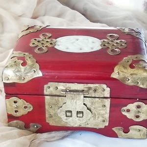 May include: A red wooden box with ornate brass corner accents and a central clasp. The box features a white oval inlay and decorative brass hardware, showcasing an Asian-inspired design. The box is likely used for jewelry or keepsakes.