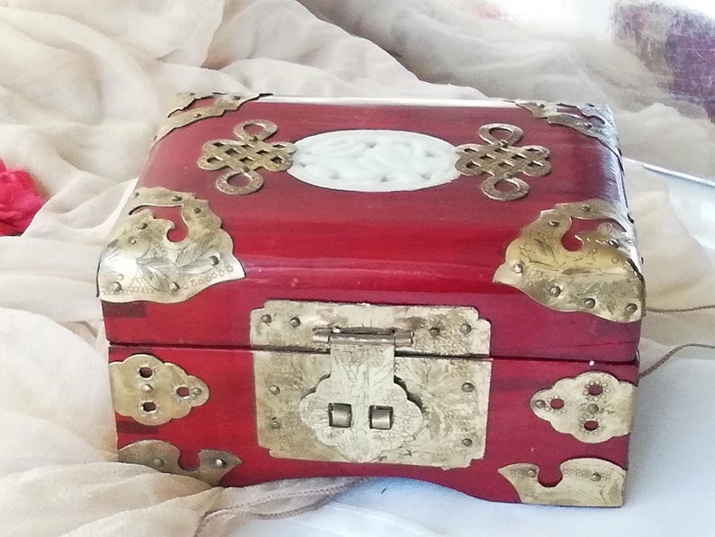 May include: A red wooden box with ornate brass corner accents and a central clasp. The box features a white oval inlay and decorative brass hardware, showcasing an Asian-inspired design. The box is likely used for jewelry or keepsakes.
