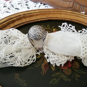 May include: A silver ring with a round, textured top. The top of the ring is covered in small, raised circles.