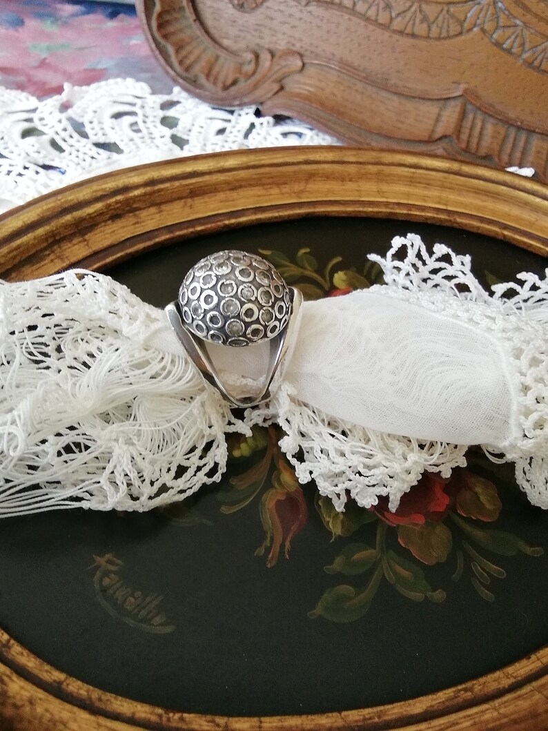 May include: A silver ring with a round, textured top. The top of the ring is covered in small, raised circles.