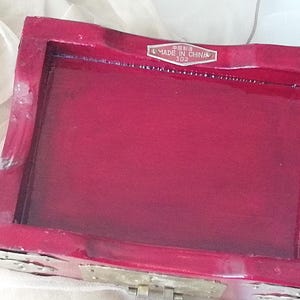 May include: A rectangular, red lacquered wooden box with a hinged lid. The box has a glossy finish and a label that reads "MADE IN CHINA". The box is likely used for jewelry or small keepsakes.