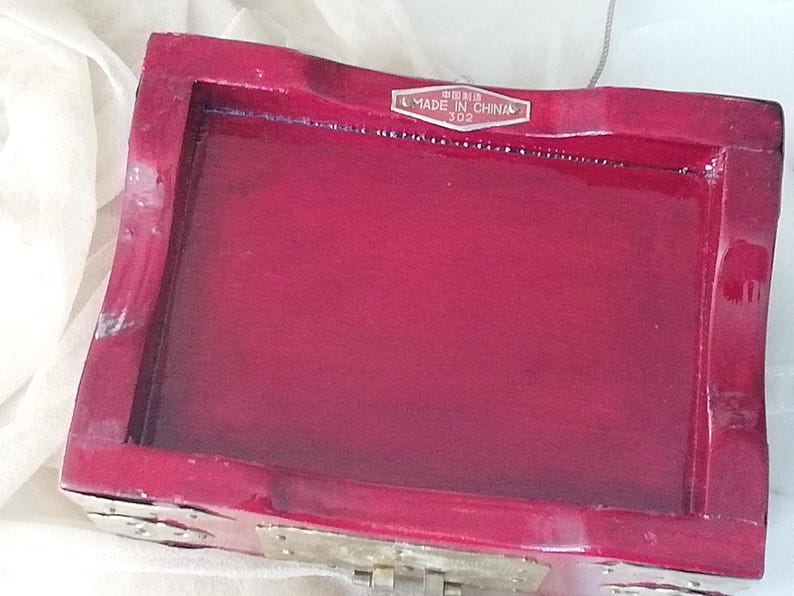 May include: A rectangular, red lacquered wooden box with a hinged lid. The box has a glossy finish and a label that reads "MADE IN CHINA". The box is likely used for jewelry or small keepsakes.