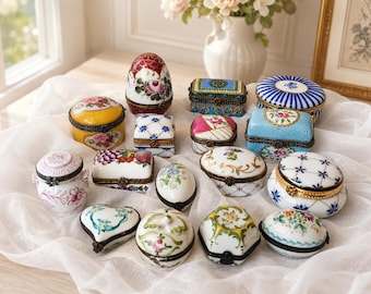 May include: A collection of fifteen small porcelain trinket boxes featuring various shapes including egg, heart, oval, and rectangular forms. Each box is decorated with hand-painted floral patterns, geometric designs, and gold-tone metal hinges.