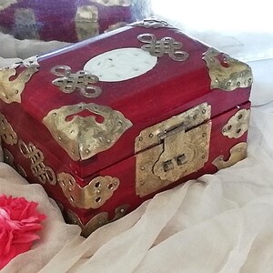May include: A red wooden box with brass corner accents and a brass clasp. The box features decorative brass embellishments and a white oval inlay on the lid. A pink flower rests near the box.
