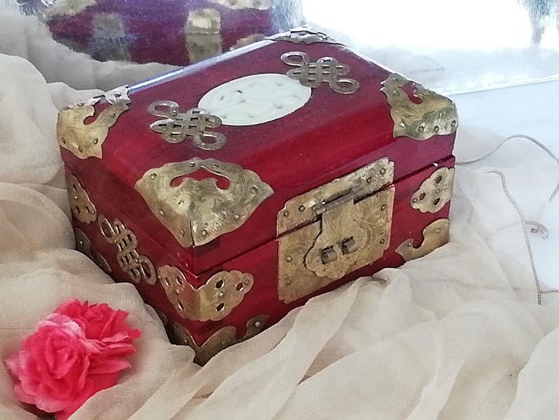 May include: A red wooden box with brass corner accents and a brass clasp. The box features decorative brass embellishments and a white oval inlay on the lid. A pink flower rests near the box.