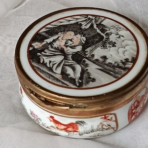May include: A small, round, antique porcelain trinket box with a hinged lid. The lid features a black and white illustration of two figures embracing. The box has a gold-colored metal rim and base, with painted floral and rooster motifs.