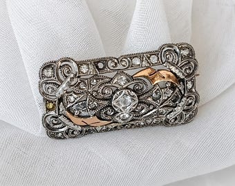 May include: A rectangular brooch featuring intricate silver-toned filigree metalwork, embedded with numerous clear sparkling stones. The design includes two smooth, curved rose gold-toned accents positioned diagonally across the center.