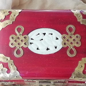 May include: A red wooden box with brass corner accents and decorative metalwork. The box features a white oval centerpiece with a carved design and two decorative metal ornaments. A vintage-style jewelry box.
