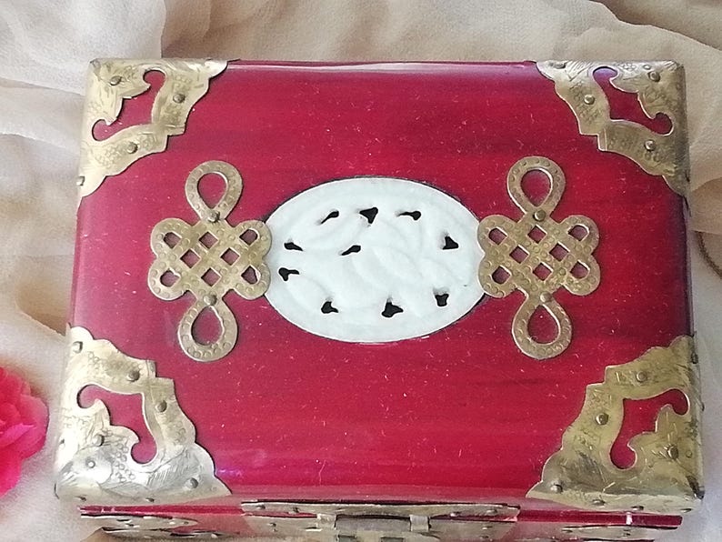 May include: A red wooden box with brass corner accents and decorative metalwork. The box features a white oval centerpiece with a carved design and two decorative metal ornaments. A vintage-style jewelry box.