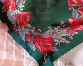 May include: A square, dark green scarf featuring a floral design. Large red poppies with detailed petals and green and blue leaves are arranged around the edges. The scarf is made of a soft, flowing fabric.