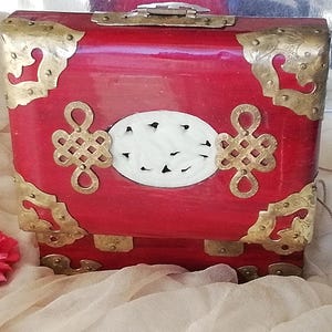 May include: A red lacquered box with brass corner accents and decorative hardware. The box features a white oval centerpiece with black accents and ornate gold-colored clasps. A red flower is visible in the lower left corner.