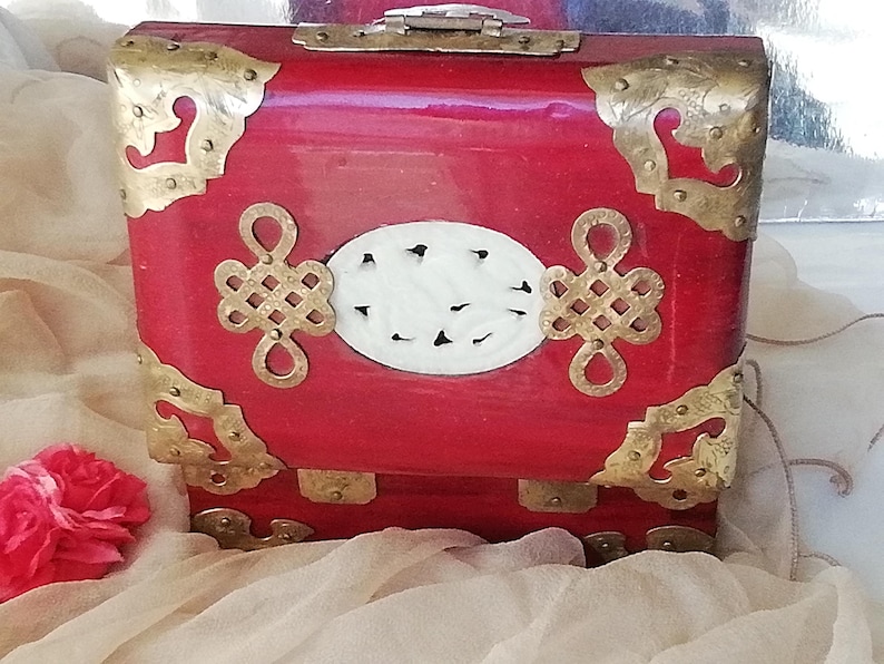 May include: A red lacquered box with brass corner accents and decorative hardware. The box features a white oval centerpiece with black accents and ornate gold-colored clasps. A red flower is visible in the lower left corner.